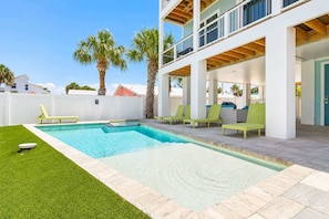 Un área de piscina moderna con piscina rectangular, césped verde y un edificio de varios niveles con balcones.