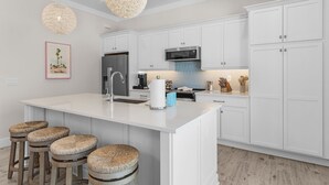 A modern kitchen with white cabinetry, a central island with stools, a stainless steel refrigerator, and a hanging light fixture. - Cabana Club · 4 Kings · 3 Min Beach (Fort Myers Beach)