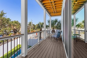 A wooden deck with a covered seating area, white railings, and a view of a swimming pool and greenery.