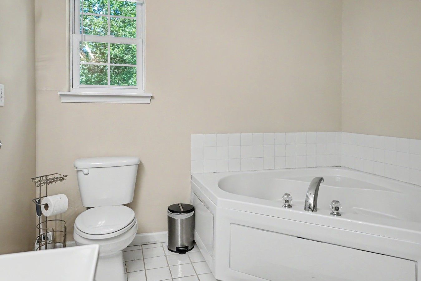 A bathroom with a toilet, a trash can, a bathtub, and a window with a view of trees.