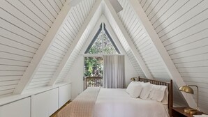 A bedroom with a sloped ceiling, a bed with white linens, a nightstand with a lamp, and a view of greenery outside. - 3BR retreat w/ fireplace & BBQ grill (Lake Arrowhead)