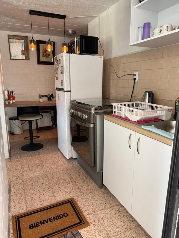 Une cuisine compacte équipée d’une cuisinière à gaz, d’un four à micro-ondes, d’un réfrigérateur et d’un évier. Un petit coin repas est aménagé avec une chaise et une table.