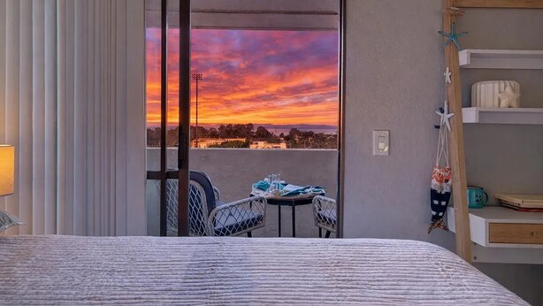 A bedroom with a bed, a small table, chairs, a balcony with a view of a sunset, and a shelf with decorative items. - Magical Monterey Carmel Pebble Beach travel, family and pet friendly whole Condo (Monterey)