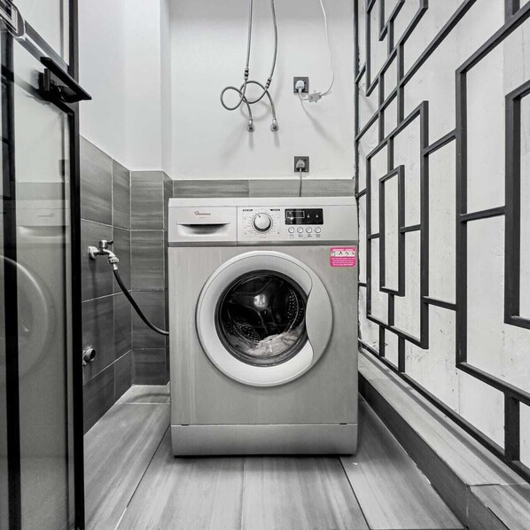 A compact, modern laundry room with a front-loading washing machine, a metal rack, and a wall-mounted electrical outlet.