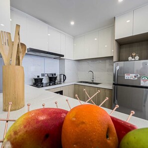Une cuisine moderne avec des meubles blancs, des appareils en acier inoxydable et un panier de fruits contenant des pommes et des oranges.