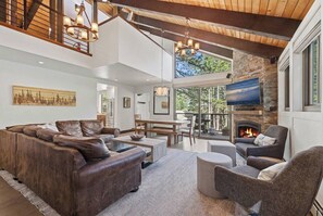 A spacious living room with a leather sectional sofa, a gray armchair, a fireplace, and a large TV mounted on a stone wall. - Tahoe City Chalet | Sleeps 10 | Summer Beach Club, Hot Tub & Sauna (Tahoe City)
