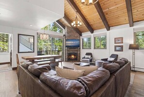 A spacious living room with a stone fireplace, leather sofas, and large windows offering a view of the outdoors. - Tahoe City Chalet | Sleeps 10 | Summer Beach Club, Hot Tub & Sauna (Tahoe City)