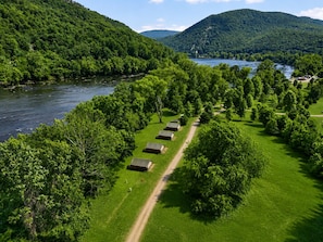 Um camping com tendas dispostas em fileiras ao longo de um caminho de terra, cercado por vegetação exuberante e um rio.