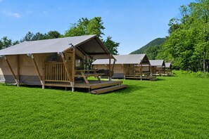 A row of wooden cabins with canvas roofs, set on a grassy field with trees and hills in the background. - Luxury Safari Tent on the New River (Blacksburg)