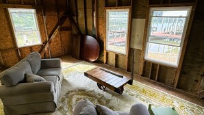 A rustic cabin interior with a sofa, a small table, and a view of a dock through large windows. - LITTLE WICOMICO WATERFRONT COMPOUND WITH KAYAKS, ARCADE, DOCKS, WESTERN SUNSETS (Reedville)