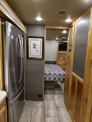 A compact RV interior with a stainless steel refrigerator, a bed with patterned bedding, a mounted television, and wooden cabinetry.