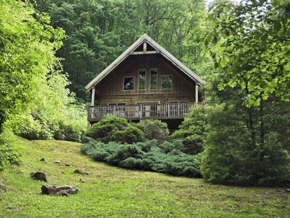 Une cabane en bois dotée d’un balcon, entourée d’une végétation luxuriante et d’une colline herbeuse.