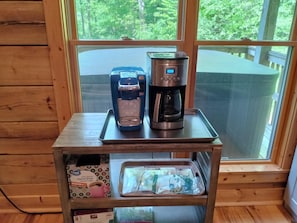 A coffee maker and a Keurig on a wooden table near a window with a view of trees.
