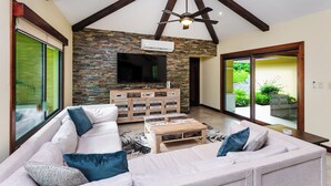 A living room with a large sofa, a coffee table, a flat-screen TV, and a stone wall. - Casa Quetzal, a charming tropical retreat in Hacienda Pinilla Beach Resort (Santa Cruz)