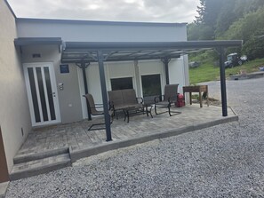 Et overdækket udendørs siddeområde med bænk, bord, en dør og en grussti.
