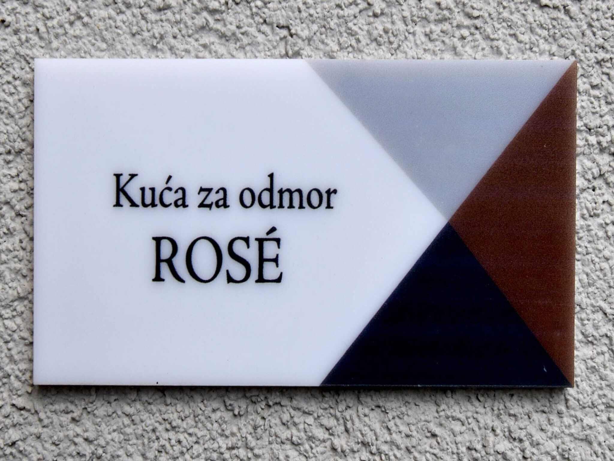 Segno bianco con testo nero che riporta 'Kuća za odmor ROSÉ' su una parete strutturata.