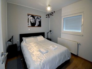 A neatly made bed with white linens, a bedside lamp, a wall-mounted picture, and a window with blinds.