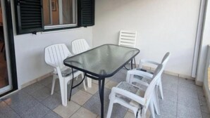 A small patio area with a glass table and white plastic chairs, a window with shutters, and a tiled floor. - Two bedroom apartment (Rogoznica)
