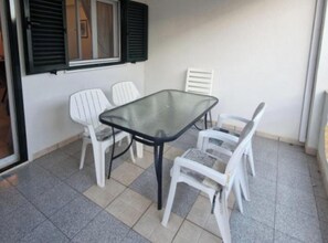 Ein kleiner Innenhof mit einem Glastisch und weißen Kunststoffstühlen, einem Fenster mit Rollläden und einem Fliesenboden.