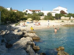 A rocky beach with clear water, people sunbathing, and white buildings in the background. - Two bedroom apartment (Rogoznica)