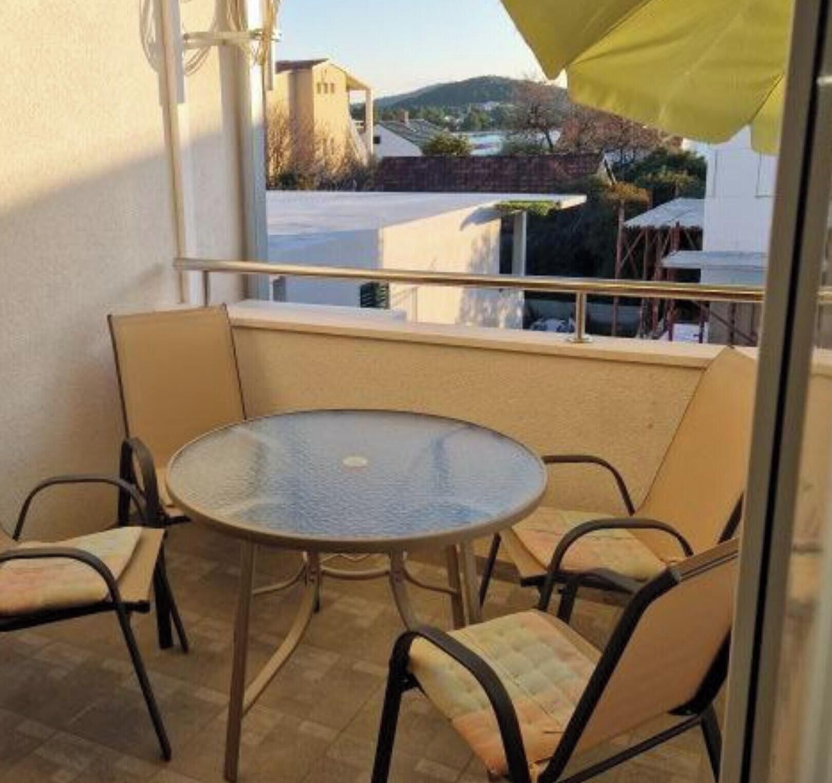 A balcony with a round glass table and four chairs, a yellow umbrella, and a view of buildings and hills.