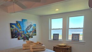 Une chambre avec un lit, un ventilateur de plafond, des œuvres d’art, une vue sur la plage et une petite table d’appoint avec un bol.