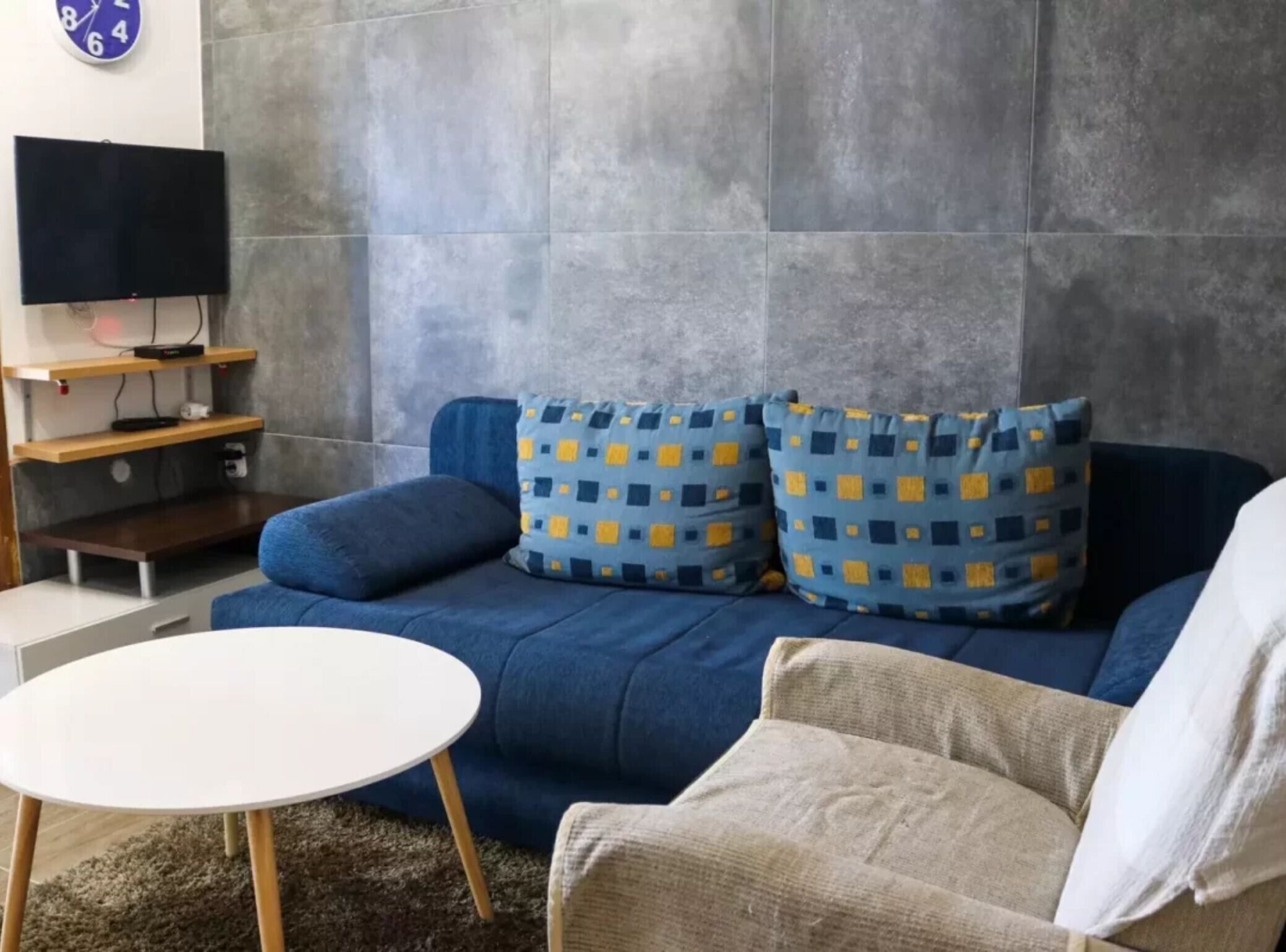 A living room with a blue sofa, patterned cushions, a white coffee table, and a flat-screen TV mounted on the wall.