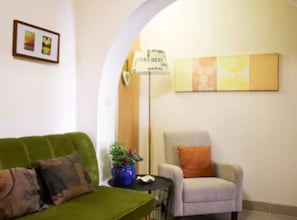 Ein Wohnzimmer mit einem grünen Sofa, einem kleinen Tisch mit einer Pflanze, einem Sessel mit einem orangefarbenen Kissen und einem Bild an der Wand.