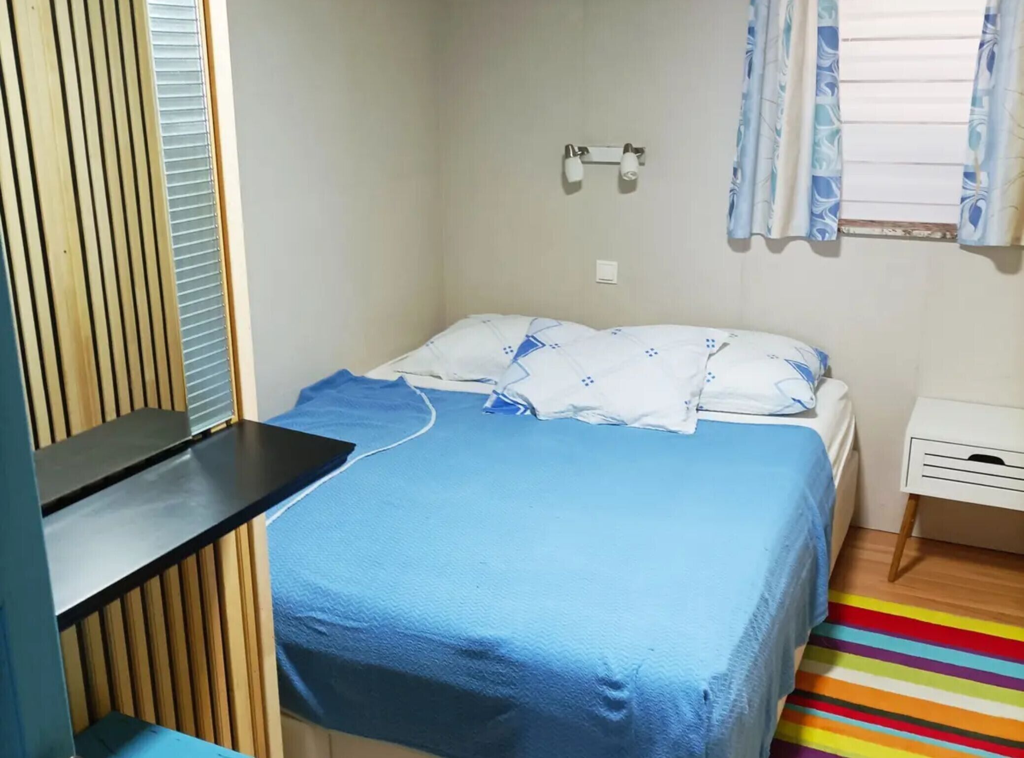 Een kleine kamer met een enkel bed, een blauw beddegoed, een houten verstelbaar raam, een witte nachtkastje en een gestreept vloerkleed.