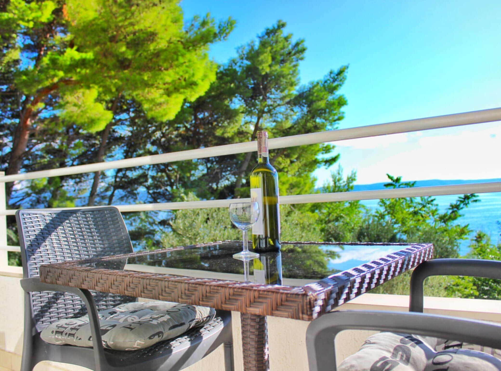 A glass table with wicker chairs, a bottle, and a glass on a balcony with a view of trees and the sea.