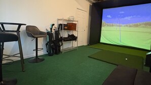 Ein Zimmer mit grünem Teppich, einer Couch, einem Barhocker, einem Golfbagständer und einer Projektionsleinwand, die einen Golfplatz zeigt.