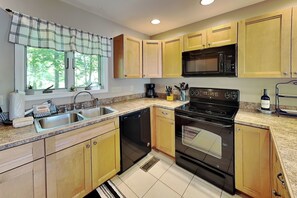 A kitchen with wooden cabinets, a black stove, a microwave, a dishwasher, and a window with curtains. - The Fairway Retreat (Oakland)