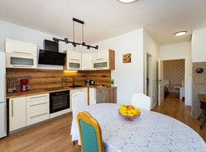 A modern kitchen with wooden cabinets, a dining table with a fruit bowl, and a hallway leading to a bedroom. - Comfort Two Bedroom Apartment (Pero) (Ploce)