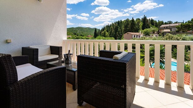 Een balkon met rieten meubels, een glazen tafel en uitzicht op een zwembad en groen.