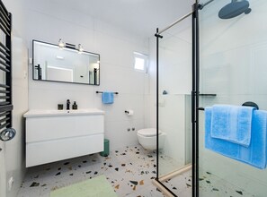 Ein Badezimmer mit einem weißen Waschbecken, Spiegel, Toilette, einer Duschkabine aus Glas und einem Terrazzo-Boden.