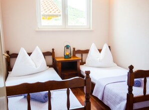 Ein Zimmer mit zwei Holzbett, weißen Kissen, einem kleinen Nachttisch mit einer Laterne und einem Fenster mit Blick auf ein Ziegeldach.