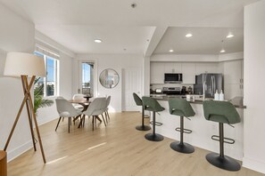 Eine moderne Küche mit weißen Schränken, einer Mikrowelle und einem Kühlschrank. Vier Barhocker und ein Esstisch mit Stühlen sind vorhanden.