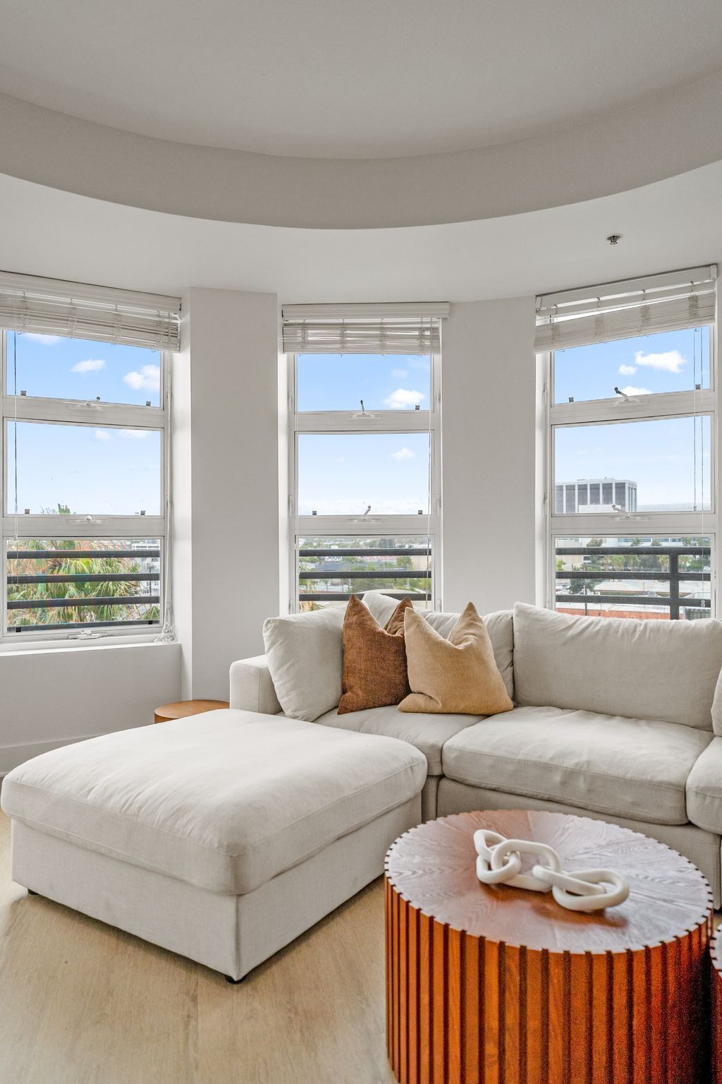 A modern living room with a sectional sofa, a round wooden coffee table, and large windows offering a city view.