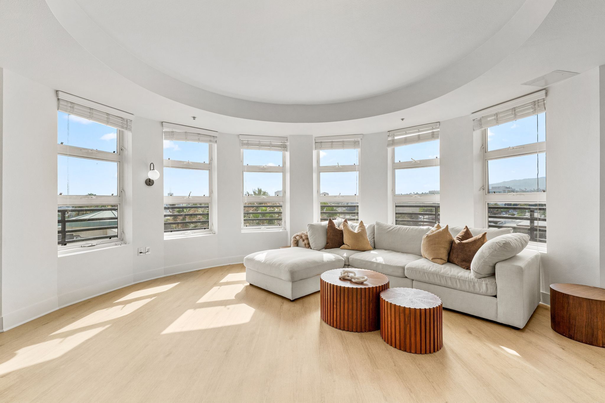 A spacious living room with a large L-shaped sofa, multiple round wooden side tables, and large windows offering a view of the city.