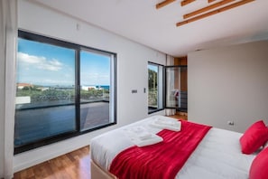 A modern bedroom with a large window, a bed with red and white bedding, and a view of the city. - Table Top by Azorean Living (Ponta Delgada, São Miguel)