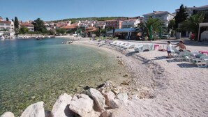 En strand med klart vand, en sandet kystlinje og bygninger i baggrunden.