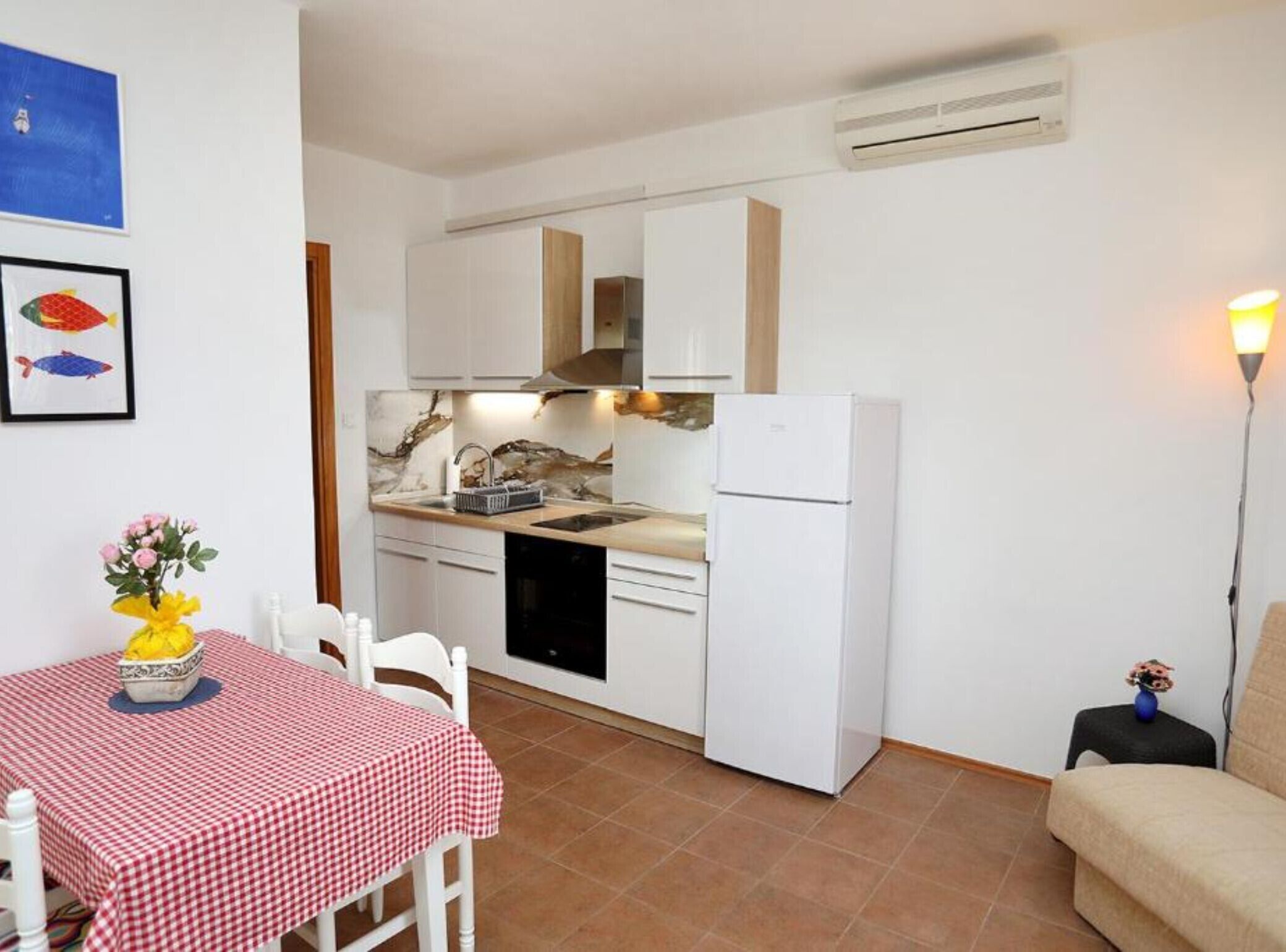 Una cucina compatta con mobili bianchi, forno integrato e frigorifero. È presente un tavolo da pranzo con tovaglia a quadri e sedie.