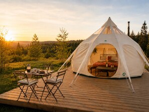Uma tenda em formato de cúpula com uma cama no interior, uma mesa com xícaras e uma panela, e cadeiras em um deck de madeira.