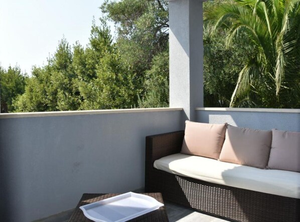Un balcon avec un canapé et des coussins en osier, une table avec un plateau, et une vue sur des arbres et un palmier.