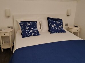 Ein Bett mit weißen Leinen und zwei blumigen, blauen Kissen, ein Nachttisch mit Fernbedienung und eine wandmontierte Lampe.