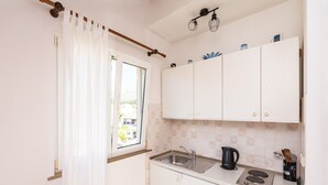 Een kleine keuken met witte kastjes, een raam met gordijn, een spoelbak, een waterkoker en een fornuis.