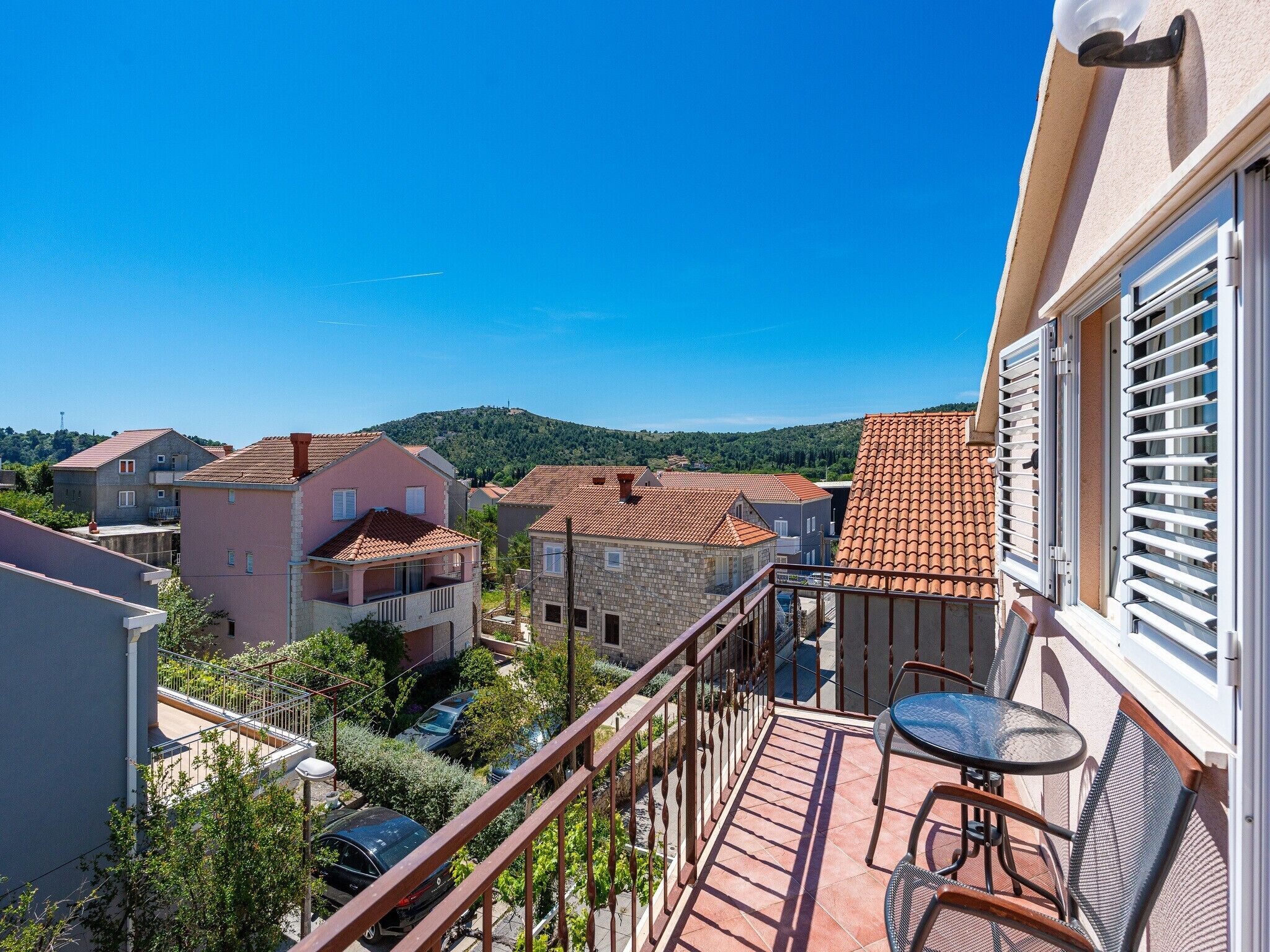 A balcony with a table and chairs, overlooking a residential area with terracotta-roofed houses and a clear blue sky.