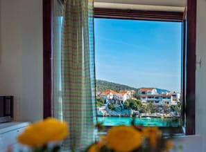 Ein Zimmer mit Blick auf eine Küstenstadt, ein Fenster mit Vorhängen und eine Vase mit gelben Blumen.