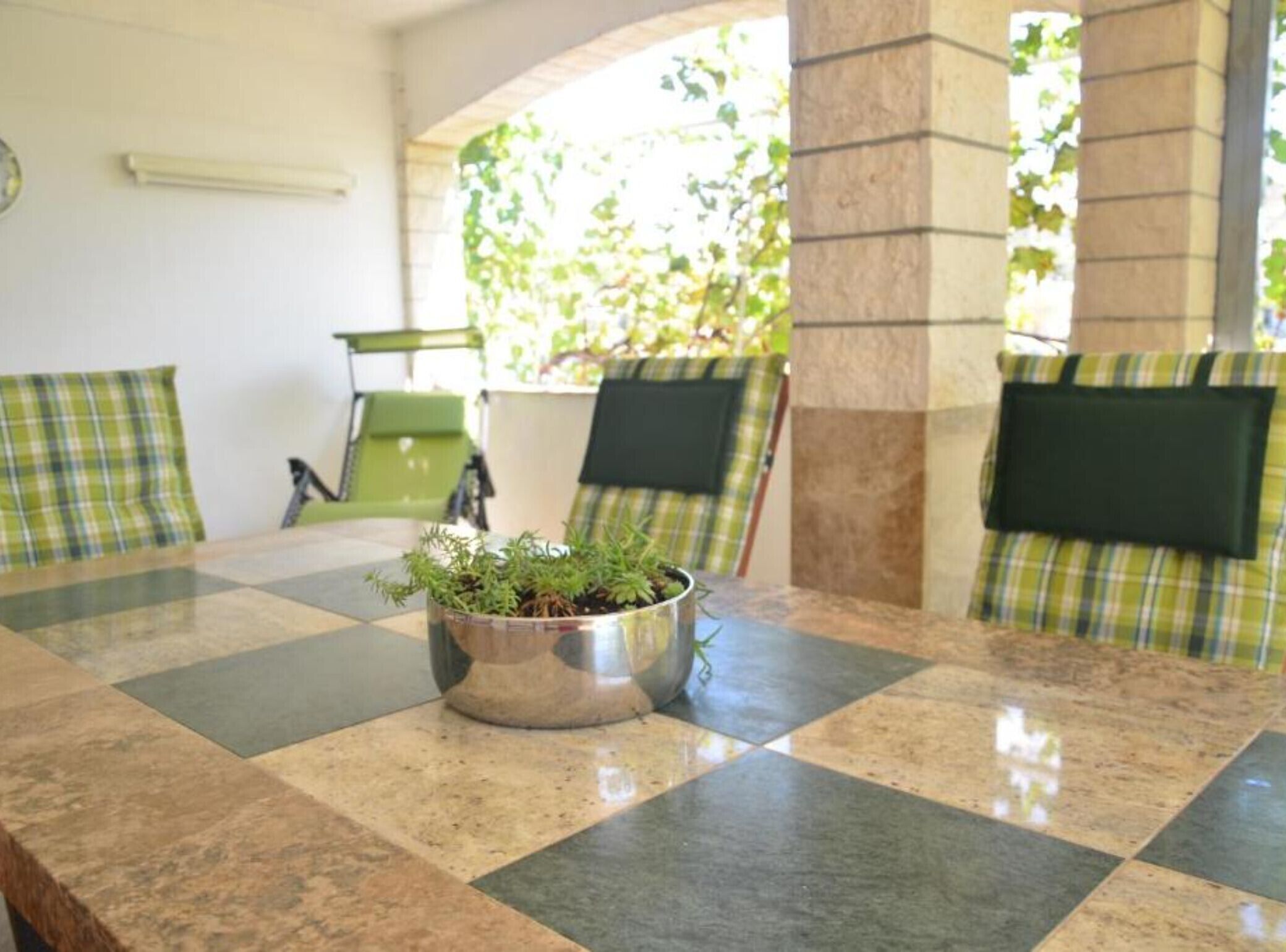 Une table à carreaux, un bol en métal rempli de plantes, des chaises vertes avec des coussins à carreaux et un pilier en pierre.