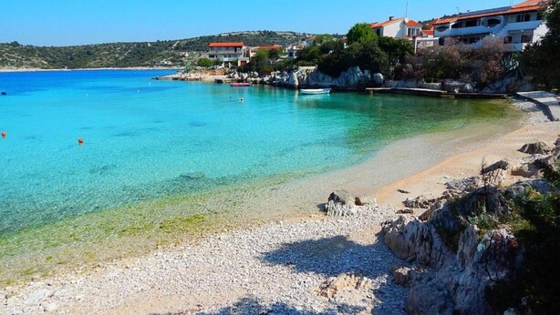 Una platja costanera amb aigua turquesa cristal·lina, sorra de còdol i edificis amb teulades vermelles.